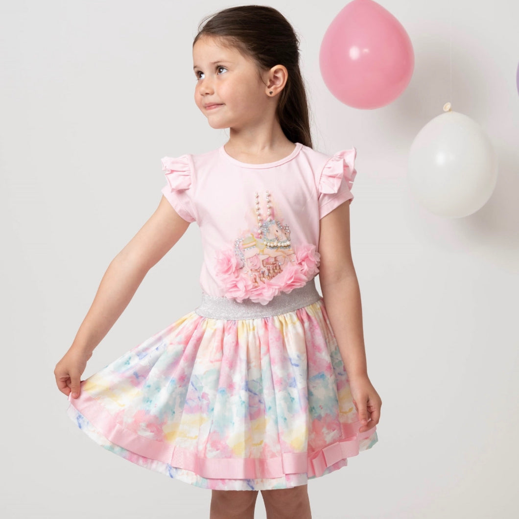 Picture of a girl wearing a Caramelo skirt set in pink which has an elasticated floral skirt with matching pink shirt sleeve tee shirt with motif of carousel horses with pearl detailing 